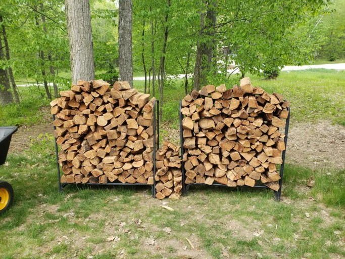 Two loaded metal firewood racks stacked on grass, with the stack weight resting on narrow rack legs